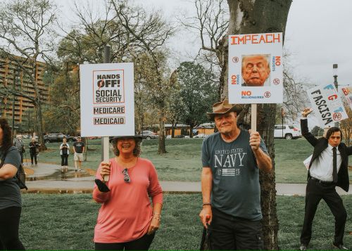 Demonstrators standing in a park holding protest signs, including “HANDS OFF! SOCIAL SECURITY MEDICARE MEDICAID,” “IMPEACH,” and “NO FASCISM,” embodying the power of protest in a peaceful public gathering.