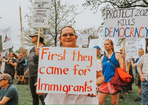 Protesters hold signs during a demonstration, including one that reads "First they came for Immigrants," highlighting public resistance to the rise of tyranny.