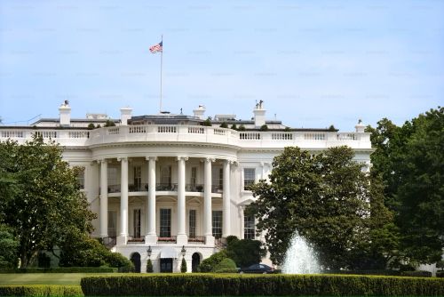 The White House on a clear summer day, with lush green trees and grass in the foreground and the American flag flying atop the building.