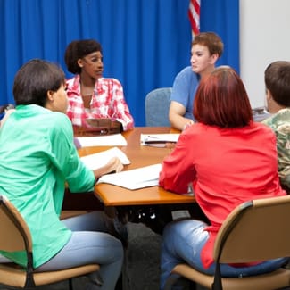 A group of students seated around a table in discussion, with notebooks, papers, and an American flag in the background. 🖋️ Caption Through dialogue and collaboration, students deepen their understanding of civic principles and democratic participation. 📜 Description This image captures a group of high school or college-aged students gathered around a rectangular classroom table, actively engaged in conversation. Notebooks, worksheets, and writing utensils are spread across the table, suggesting a structured discussion—likely related to civics, government, or U.S. history. The American flag and blue curtain in the background lend symbolic weight to the setting. The students’ focused postures and shared materials evoke a spirit of inquiry, respect, and civic learning—emphasizing the importance of dialogue in shaping informed citizens. Let me know if you'd like to adapt this for a specific age group, platform, or content theme. I can also help you create a consistent style for future visuals.