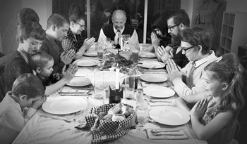 Black-and-white photo of a multi-generational family seated around a dining table, heads bowed and hands clasped in prayer before a meal. The table is set with plates, glasses, candles, and bread, capturing a solemn moment of unity and religious observance.