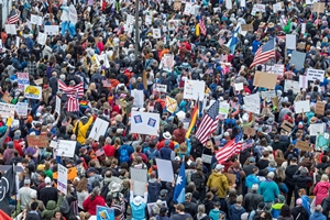 A large crowd of American protesters holding signs and banners during a No Kings demonstration, with several U.S. flags visible.